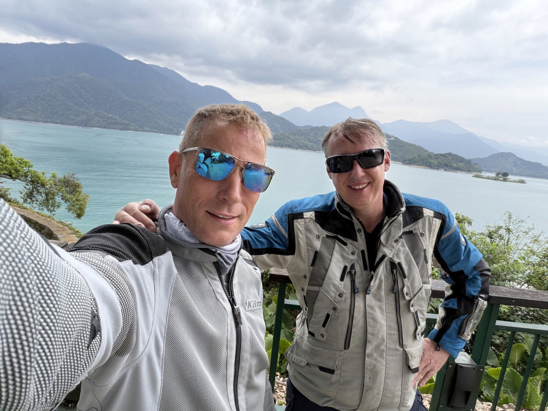 Two riders in adventure gear at a Taiwan lake with mountains behind